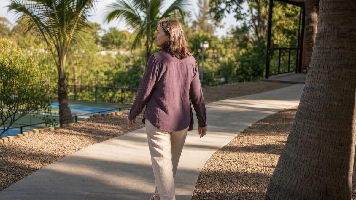 Woman walking a sunlit path through the grounds of a Chiang Mai rehab, sports court below