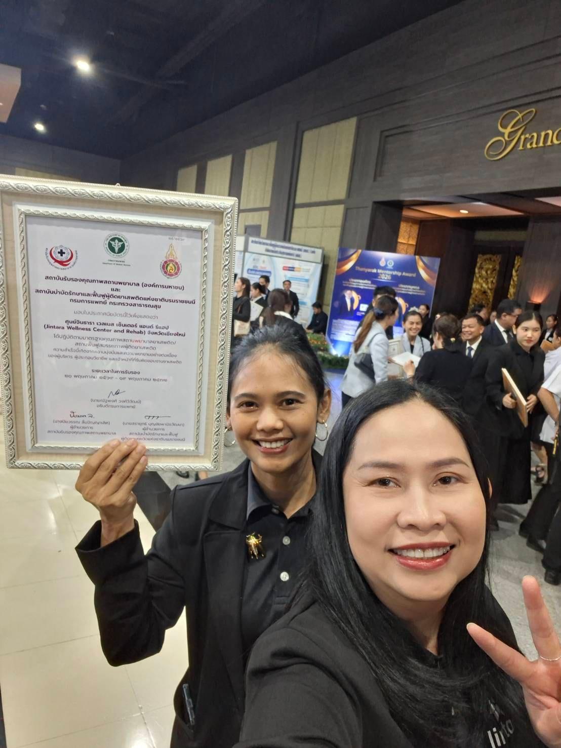 Two Jintara Rehab team members holding the framed national accreditation certificate in Thailand