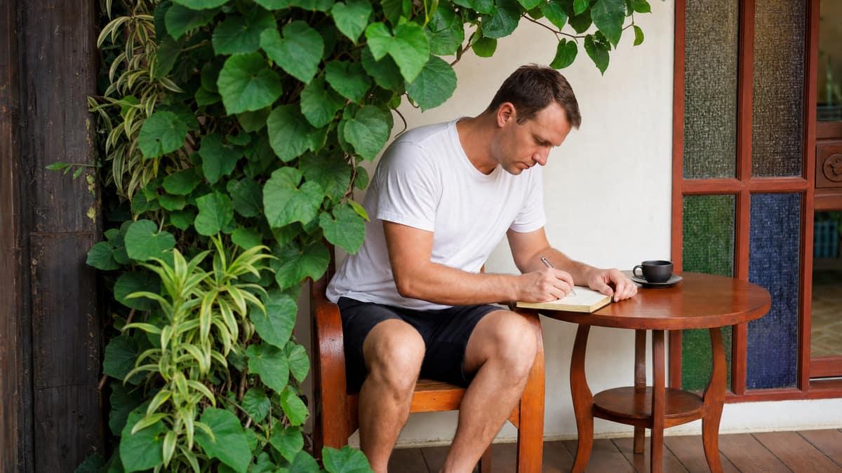 Person sitting on a wooden bench journaling on a tropical porch at Jintara Rehab Chiang Mai