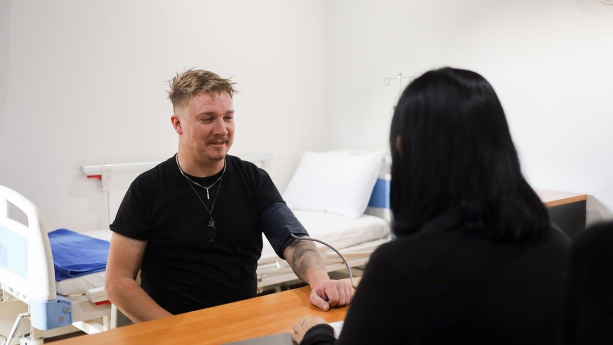 A man wearing a blood pressure cuff sits across a desk from a woman, with an examination bed behind them at Jintara Rehab