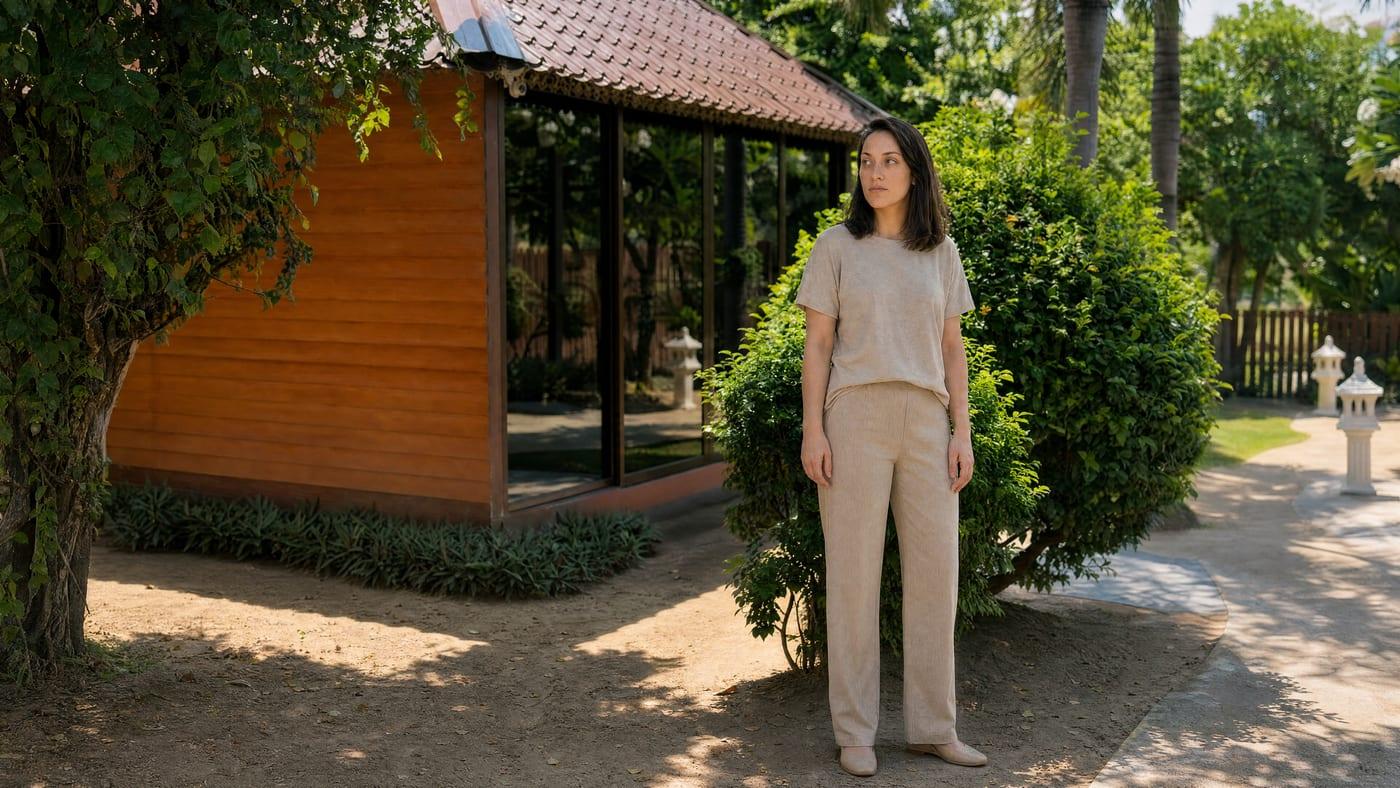 A woman stands on a sandy garden path beside a timber and glass sala in bright midday light