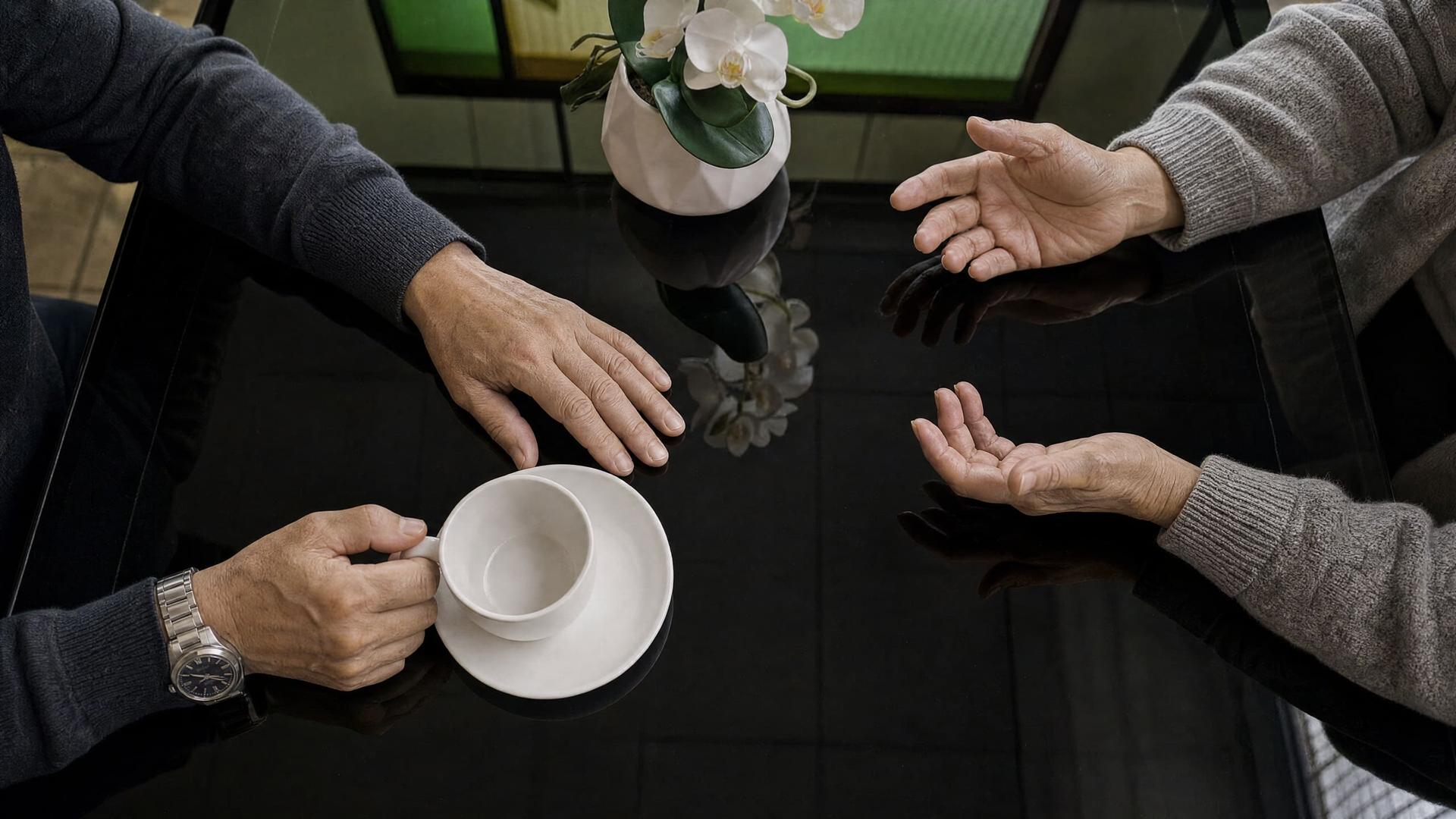 Two pairs of hands either side of a coffee cup on a glass dining table at a rehab