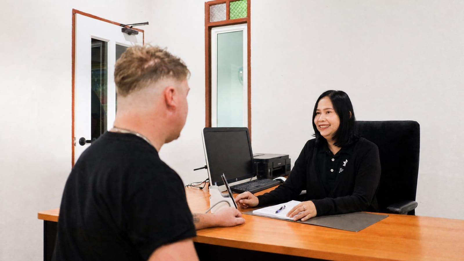 Smiling clinician in a black Jintara polo consulting a male client at a desk for polydrug assessment at Jintara Rehab Chiang Mai
