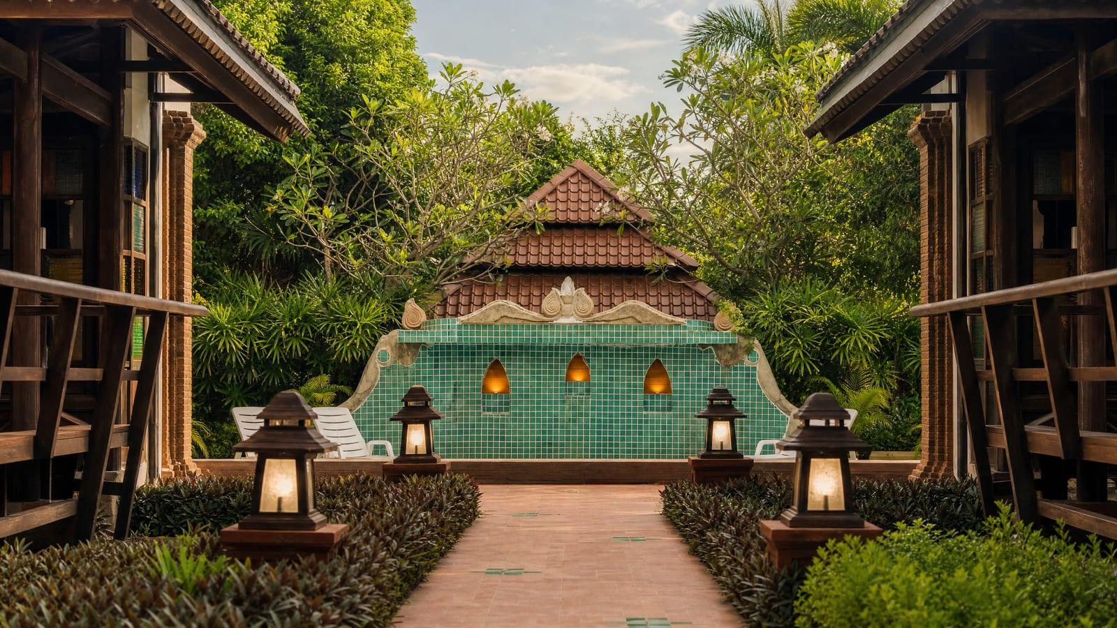 Curved garden path beside a glass-wall wooden pavilion at Jintara Rehab Chiang Mai with frangipani trees and trimmed hedges in bright morning daylight