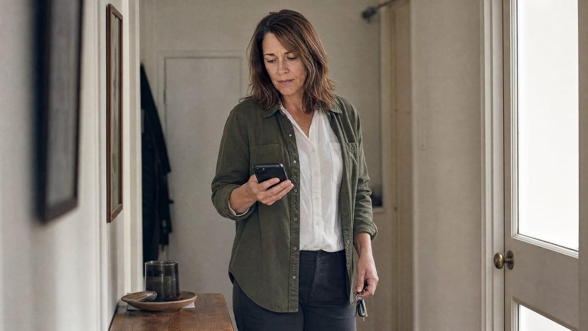 A woman in her forties stands in the hallway of her home holding her phone and her keys, deciding what to do after a family member has refused help