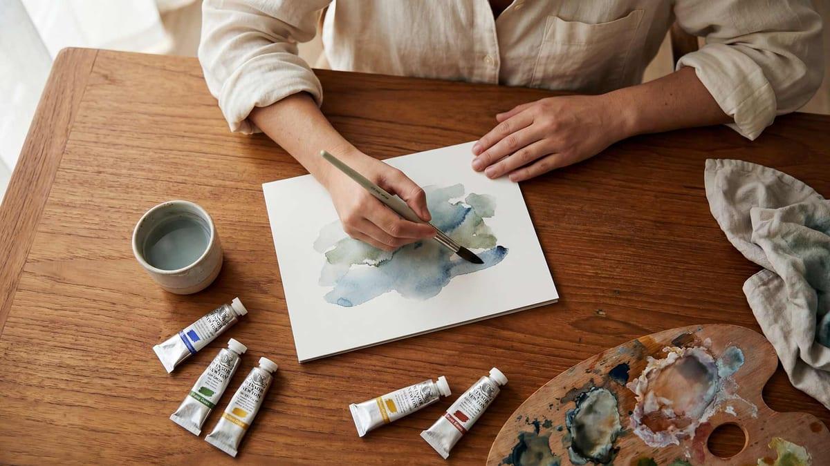Close-up of hands painting a watercolour with paints and palette on a wooden table at Jintara Rehab