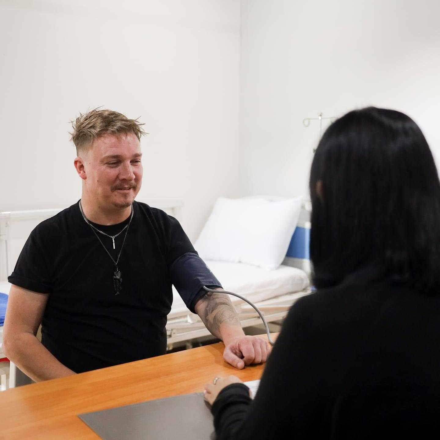A man wearing a blood pressure cuff sits across a desk from a woman, with an examination bed behind them at Jintara Rehab