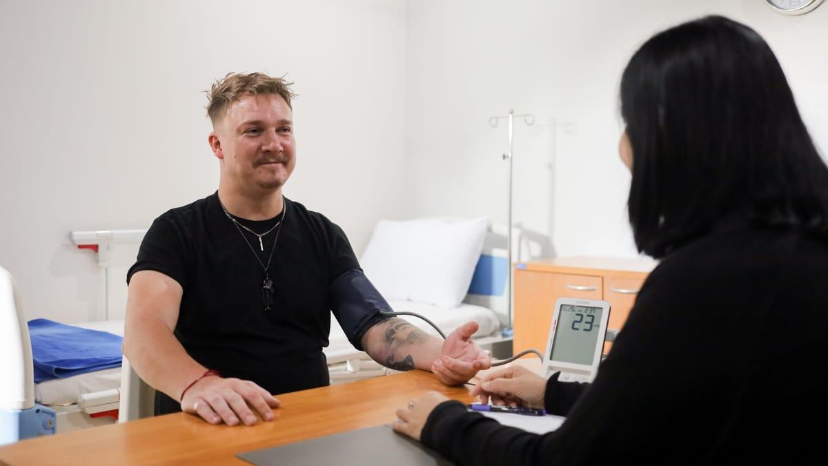 A man wearing a blood pressure cuff sits across a desk from a woman, with an examination bed behind them