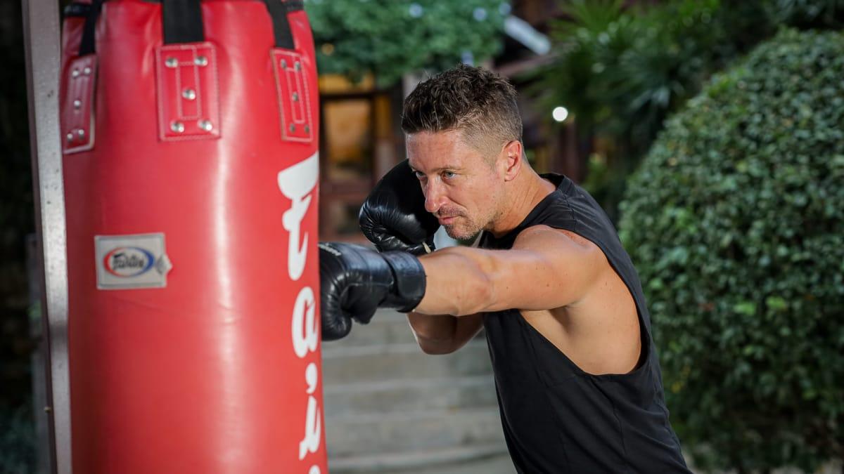 A man in boxing gloves throws a punch at a red heavy bag in the garden at Jintara Rehab in Chiang Mai