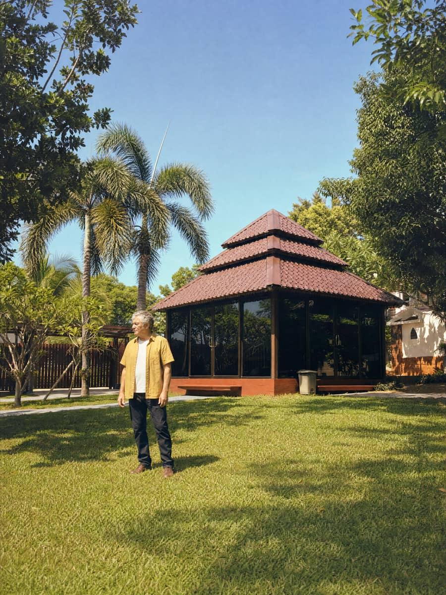 Grey-haired man standing on a wide open lawn beside a glass sala and tall palms