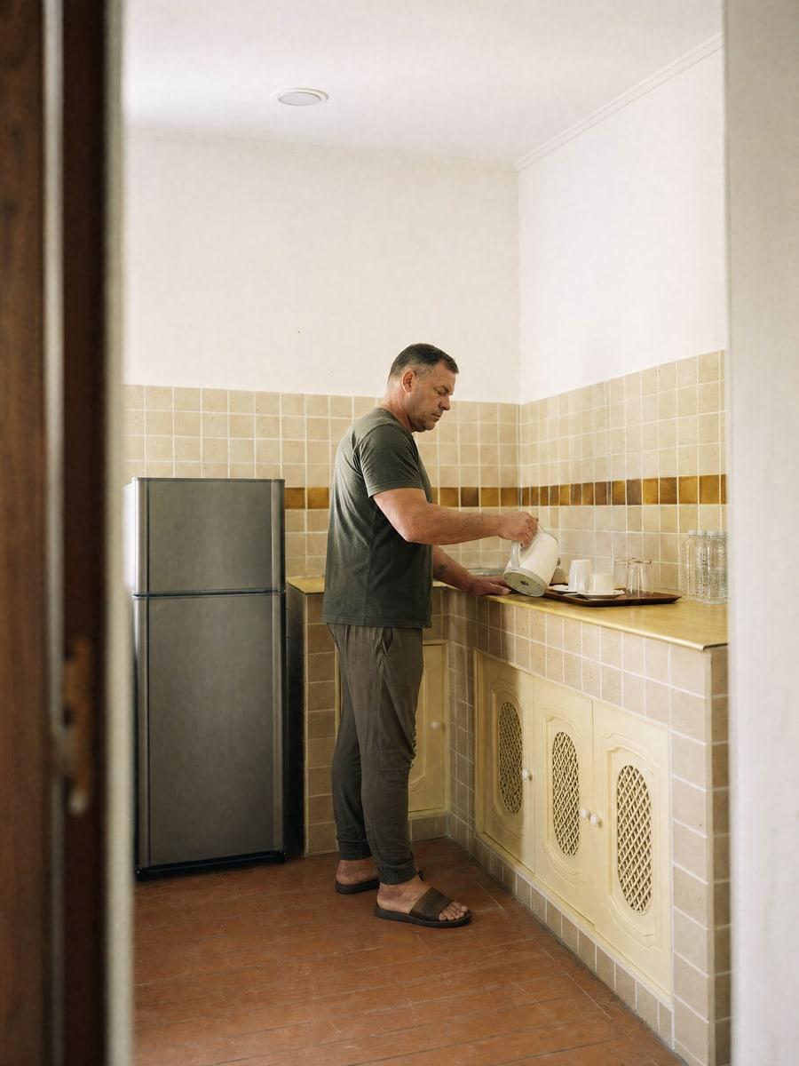 Man pouring a kettle at a tiled kitchenette counter with cups and water bottles