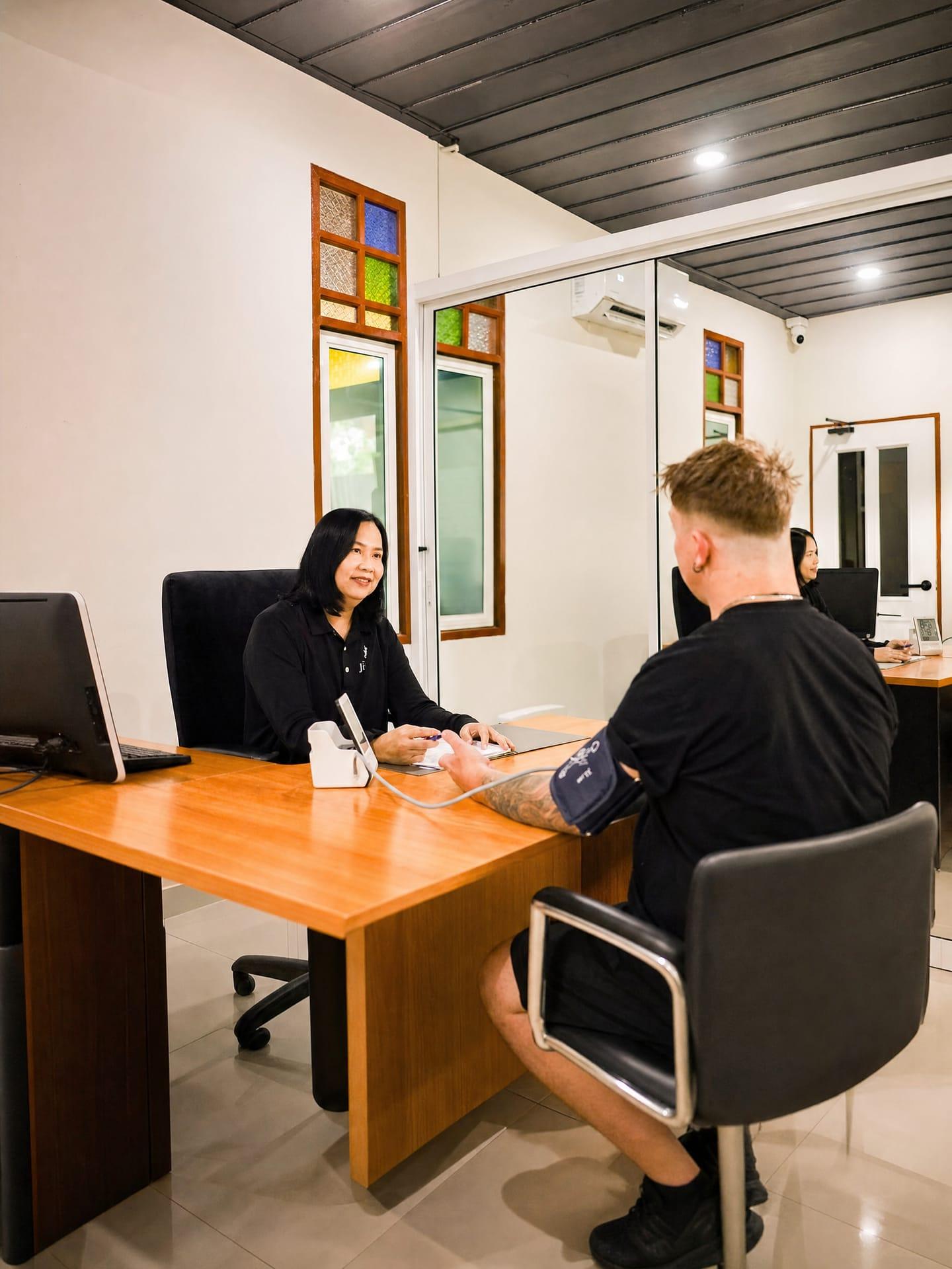 A nurse taking a blood pressure reading from a client at a timber desk in the Jintara medical office
