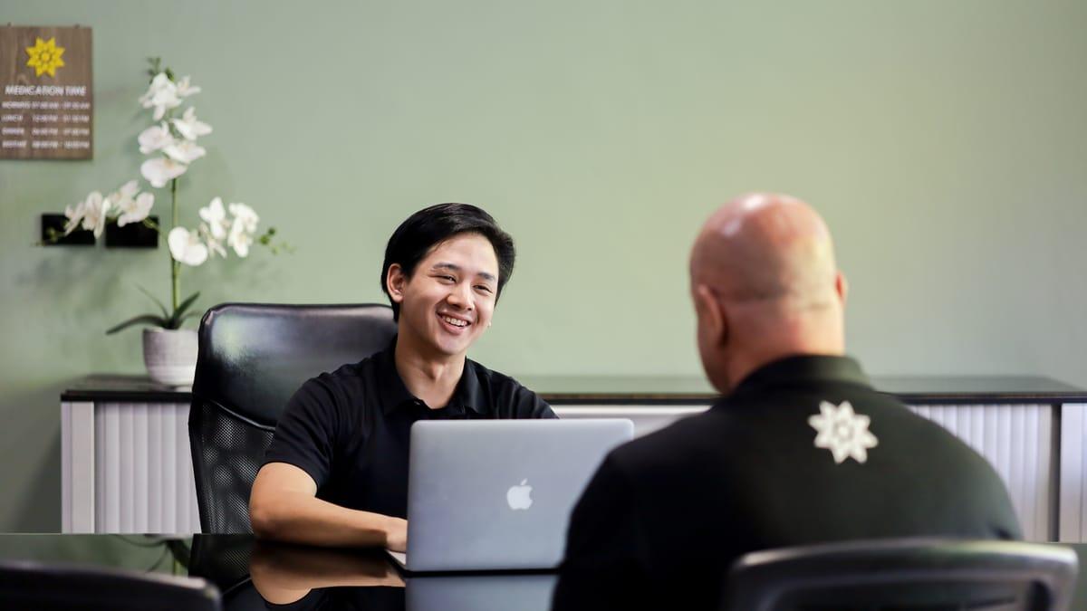 Two men talk across a desk with an open laptop and white orchids at Jintara Rehab