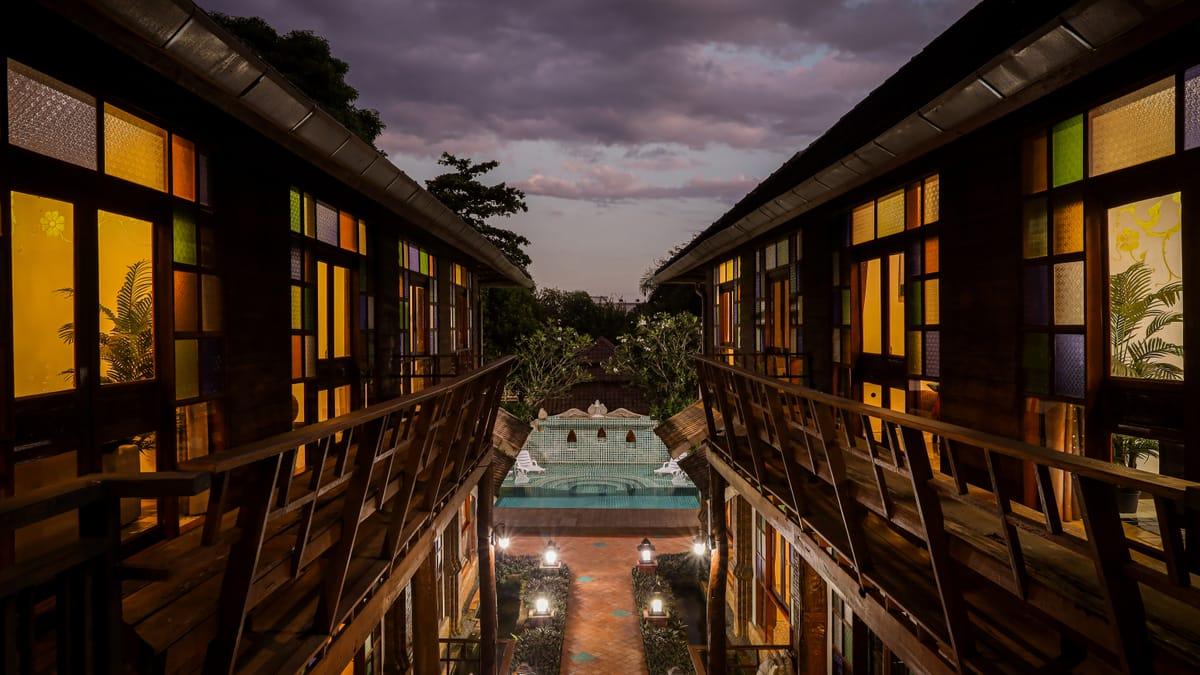Lit windows along two teak buildings at dusk, with the pool at the end of the walkway, at Jintara Rehab in Chiang Mai